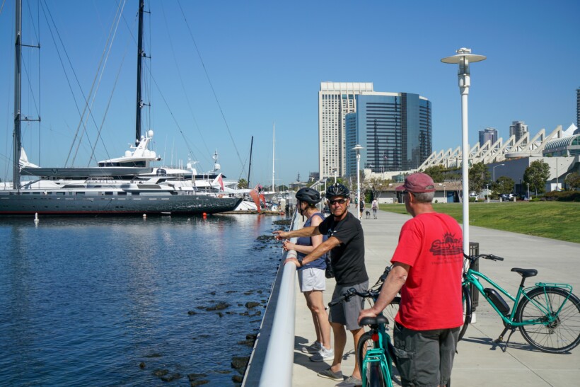 A group of cyclists stop along the water to look at the ships in San Diego
